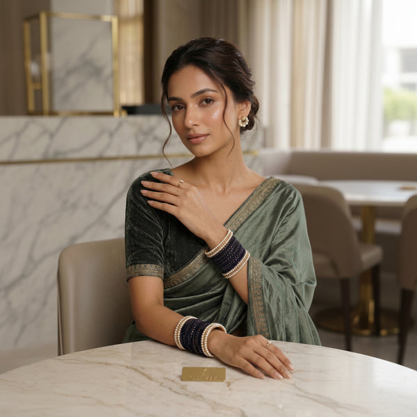 A woman wearing a sage green velvet saree with gold border detailing, paired with dark navy and cream bangles, seated indoors at a marble table.