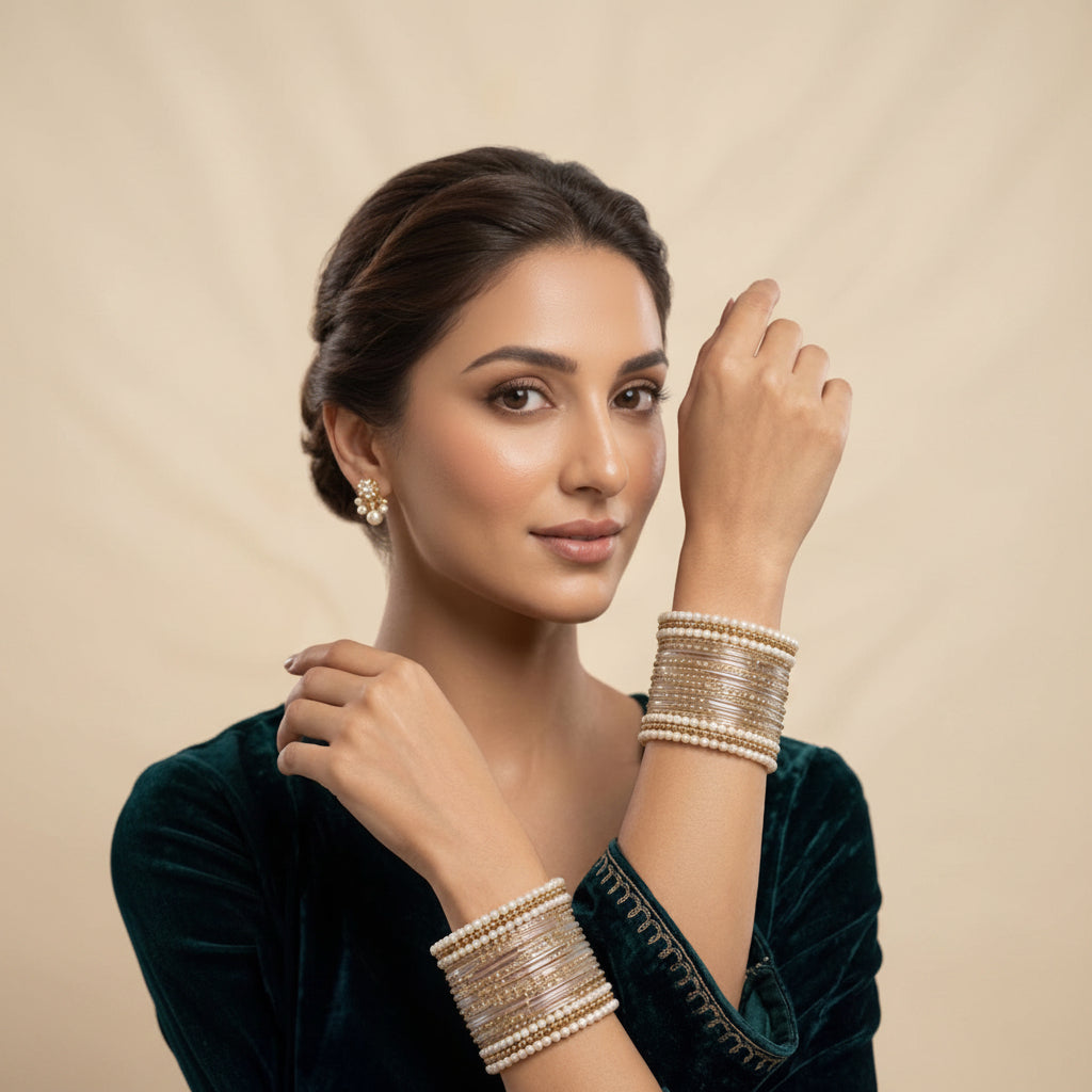 A woman wearing multiple stacked bangles on both wrists, with one bangle resting near her neck. She is wearing a dark teal velvet top against a warm beige studio background.